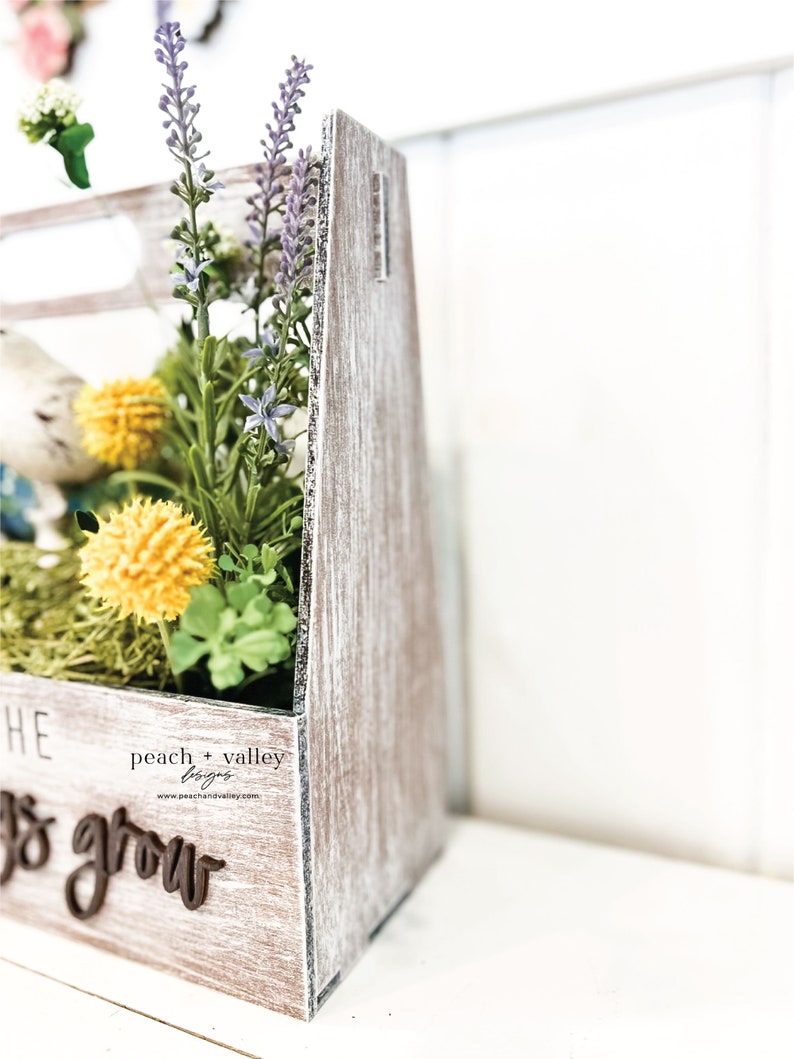 May include: A wooden crate with a whitewashed finish, filled with artificial flowers and greenery. The crate has a sign that reads "peach + valley designs" and "Let's grow".