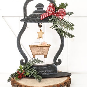 May include: A black metal lantern with a red and white bow, a star, and a manger scene hanging from it. The manger scene has the text "The gift that forever changed the world". The lantern is sitting on a wooden slice with the text "peach valley" on it.
