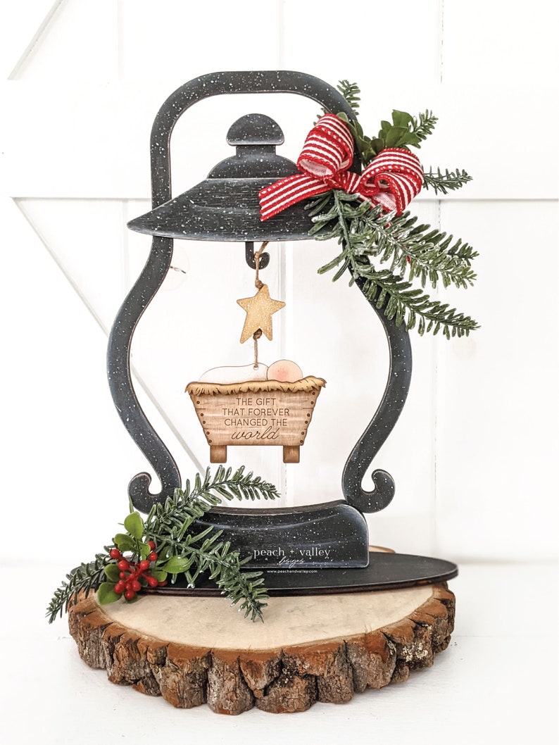 May include: A black metal lantern with a red and white bow, a star, and a manger scene hanging from it. The manger scene has the text "The gift that forever changed the world". The lantern is sitting on a wooden slice with the text "peach valley" on it.