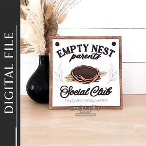Puede incluir: Un letrero de madera con el texto "Empty Nest parents Social Club" y las palabras "Empty Nest, Happy Hearts" debajo. El letrero tiene un fondo marrón y texto blanco. Hay un nido de pájaro marrón con ramitas y hojas en el letrero.
