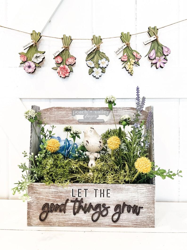 May include: A wooden crate with a whitewashed finish and the text "Let the good things grow" in black lettering. The crate is filled with artificial greenery, flowers, and a small bird figurine. A banner of wooden flower bouquets hangs above the crate.