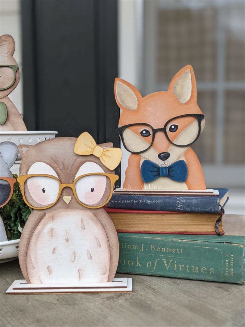 May include: A wooden owl and fox figurine with glasses and a bow tie. The figurines are standing on a stack of books with the title "Book of Virtues".