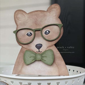 May include: Wooden bear figurine wearing green glasses and a green bow tie. The bear is painted brown and has a white background.