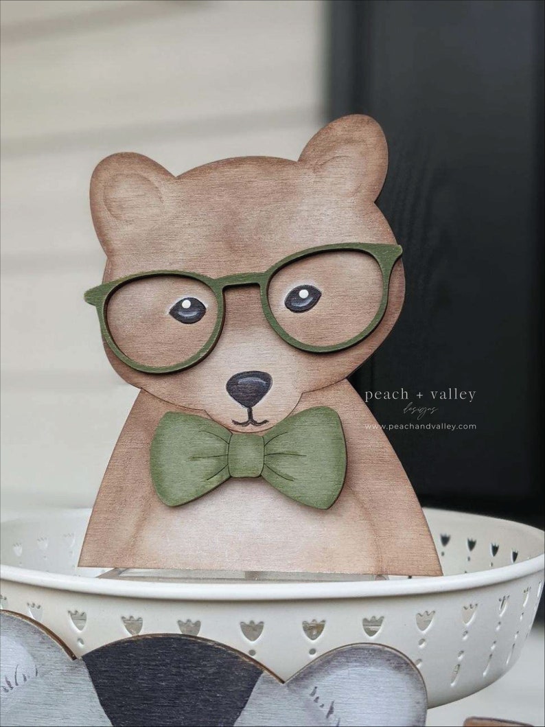 May include: Wooden bear figurine wearing green glasses and a green bow tie. The bear is painted brown and has a white background.