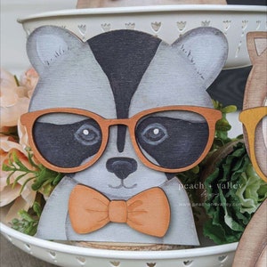 May include: A wooden raccoon figurine wearing orange-rimmed glasses and an orange bow tie. The raccoon is painted in shades of gray, black, and white. The figurine is sitting on a white tiered tray with green plants in the background. The text "peach + valley" is visible in the bottom right corner of the image.