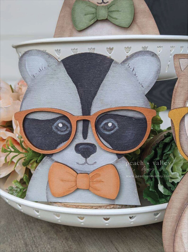 May include: A wooden raccoon figurine wearing orange-rimmed glasses and an orange bow tie. The raccoon is painted in shades of gray, black, and white. The figurine is sitting on a white tiered tray with green plants in the background. The text "peach + valley" is visible in the bottom right corner of the image.