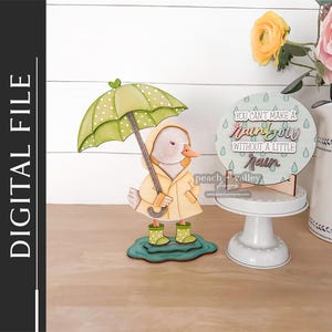 May include: A wooden duck figurine in a yellow raincoat with a green polka-dot umbrella. A round sign displays the text "You can't make a rainbow without a little rain". The theme is weather and rain-related, with a whimsical design.