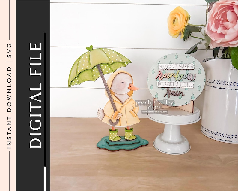 Pode incluir: Uma figura de pato de madeira usando uma capa de chuva amarela e segurando um guarda-chuva verde com bolinhas. Uma placa redonda exibe o texto "You can't make a rainbow without a little rain". O tema &eacute; clima e chuva.