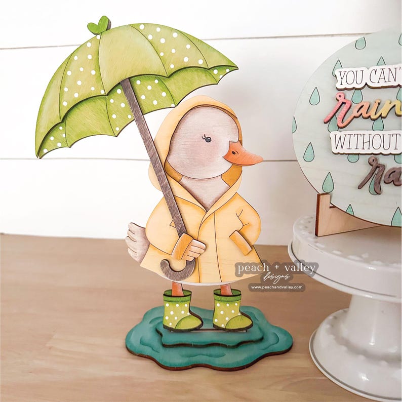 Pode incluir: Uma figura de madeira de um pato com uma capa de chuva amarela e botas de chuva verdes com bolinhas, segurando um guarda-chuva verde com bolinhas. O pato est&aacute; sobre uma base azul em forma de po&ccedil;a. Uma placa redonda com o texto "You can't have a rainbow without rain" tamb&eacute;m &eacute; vis&iacute;vel.
