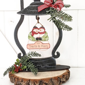 May include: A black metal lantern with a wooden base, decorated with a red and white bow, green pine sprigs, and red berries. The lantern features a cutout of two snowmen holding hands, with the text "2022 Travis & Mary" on a sign below them.