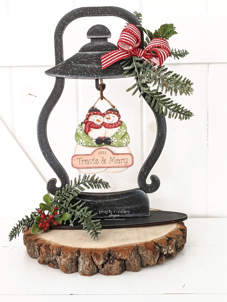 May include: A black metal lantern with a wooden base, decorated with a red and white bow, green pine sprigs, and red berries. The lantern features a cutout of two snowmen holding hands, with the text "2022 Travis & Mary" on a sign below them.