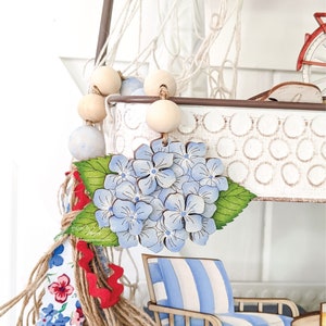 May include: A wooden hydrangea flower with blue and white painted petals, green leaves, and a brown stem. The flower is hanging from a white metal planter with a white and brown rope net. The planter is decorated with a white and brown polka dot pattern.