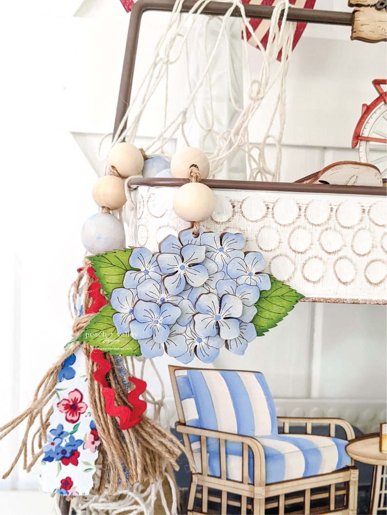 May include: A wooden hydrangea flower with blue and white painted petals, green leaves, and a brown stem. The flower is hanging from a white metal planter with a white and brown rope net. The planter is decorated with a white and brown polka dot pattern.