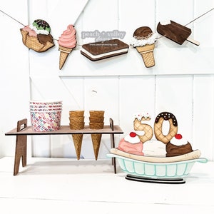 May include: Wooden ice cream cone and ice cream sandwich banner with the text "peach + valley designs" and website address.  Wooden ice cream cones and cups on a stand with a wooden banana split with the number 50 on top.