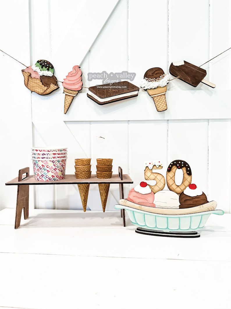 May include: Wooden ice cream cone and ice cream sandwich banner with the text "peach + valley designs" and website address.  Wooden ice cream cones and cups on a stand with a wooden banana split with the number 50 on top.