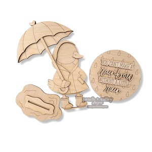 Pode incluir: Estatueta de pato de madeira com guarda-chuva, capa de chuva e galochas. Inclui tamb&eacute;m um letreiro redondo de madeira com o texto "You can't make a rainbow without a little rain" e uma po&ccedil;a de madeira. Os artigos s&atilde;o feitos de madeira clara.