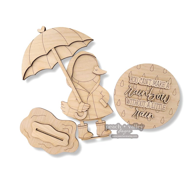 Pode incluir: Estatueta de pato de madeira com guarda-chuva, capa de chuva e galochas. Inclui tamb&eacute;m um letreiro redondo de madeira com o texto "You can't make a rainbow without a little rain" e uma po&ccedil;a de madeira. Os artigos s&atilde;o feitos de madeira clara.