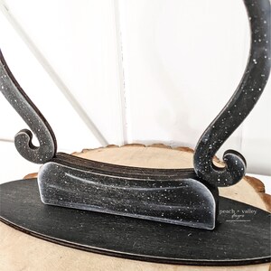 May include: Black wooden display stand with a unique curved design. The stand has a distressed finish with white speckles. The stand is sitting on a wooden surface. "peach + valley designs" is written on the stand.