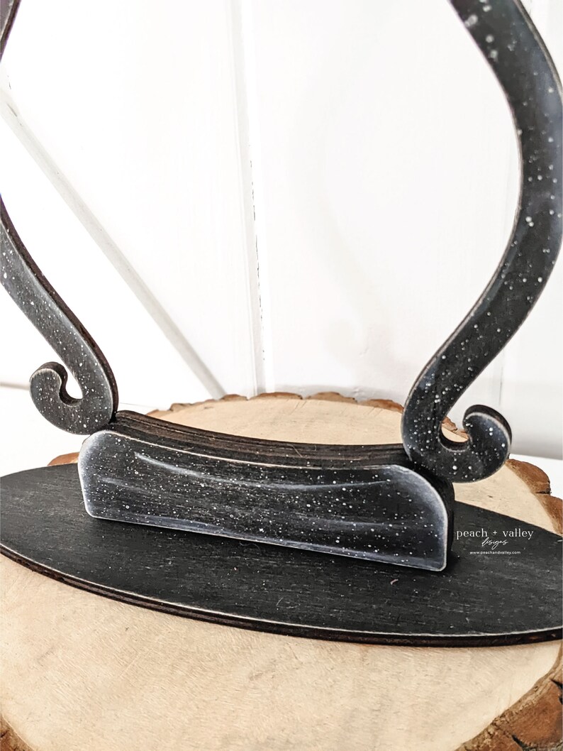 May include: Black wooden display stand with a unique curved design. The stand has a distressed finish with white speckles. The stand is sitting on a wooden surface. "peach + valley designs" is written on the stand.