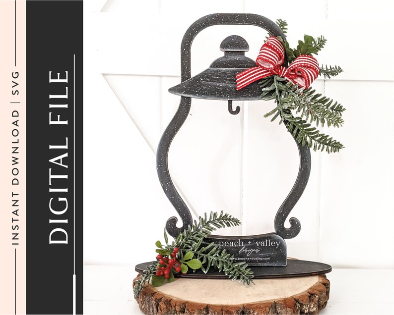 May include: Black metal lantern with a red and white striped bow and greenery. The lantern is sitting on a wooden slice with a sign that reads "peach + valley designs".