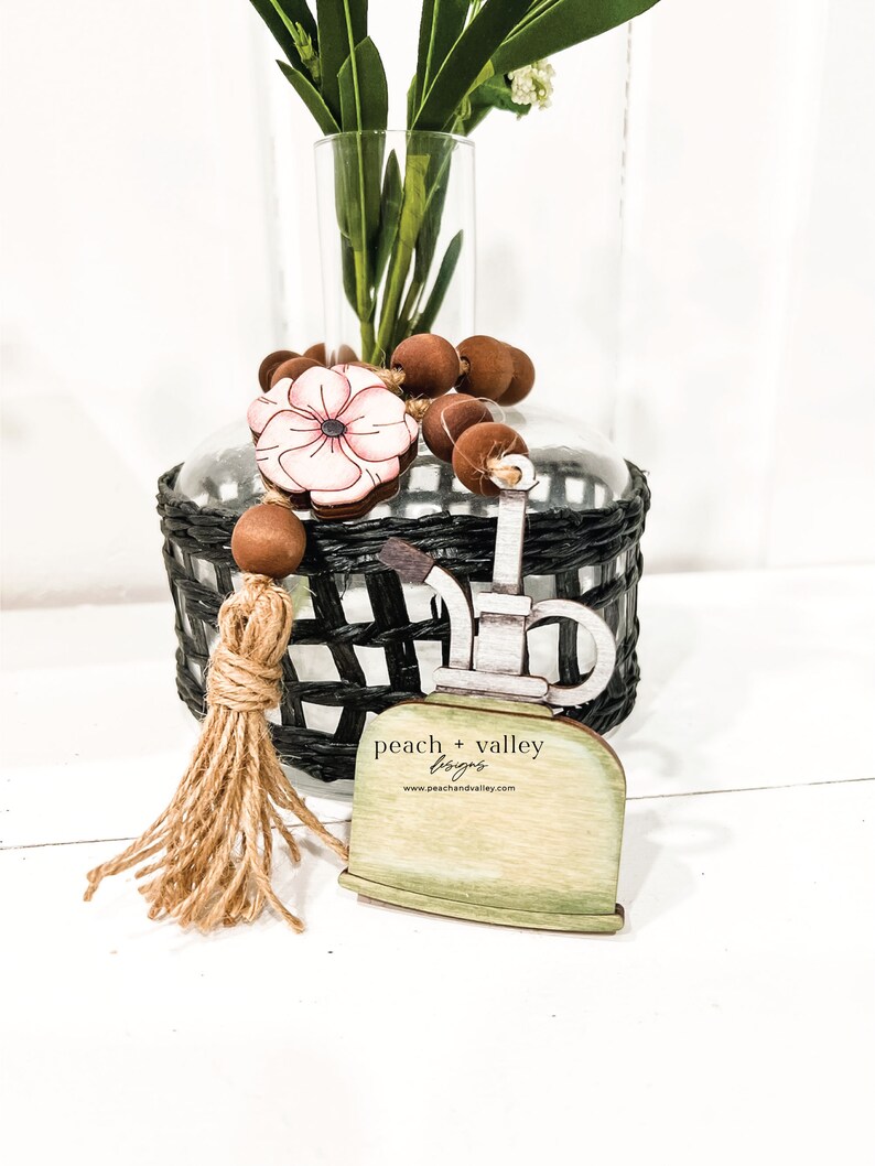 May include: Wooden farmhouse style sign with a watering can design, a pink flower, and brown beads. The sign has a tan tassel and the text "peach + valley flags" and "www.peachandvalley.com".