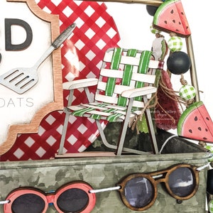 May include: A wooden sign with the text "Good Eats" and a red and white checkered background. A miniature wooden lawn chair with green and white stripes is in front of the sign. A string of watermelon and green and black beads hangs from the top of the sign. A wooden pair of sunglasses hangs from a string in front of the sign.