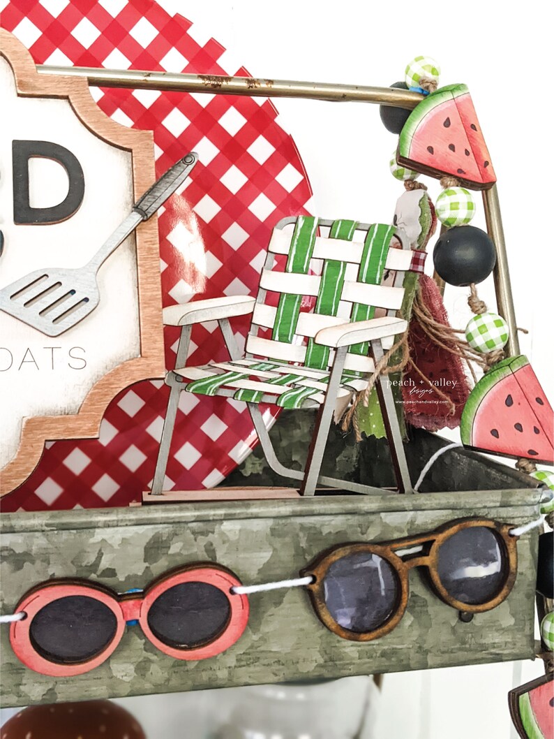 May include: A wooden sign with the text "Good Eats" and a red and white checkered background. A miniature wooden lawn chair with green and white stripes is in front of the sign. A string of watermelon and green and black beads hangs from the top of the sign. A wooden pair of sunglasses hangs from a string in front of the sign.