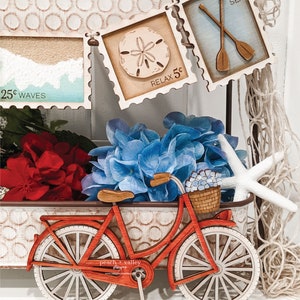May include: A red wooden bicycle with a basket filled with blue flowers. The bicycle is sitting on a white surface with a brown wooden tray behind it. The tray has a variety of wooden signs with nautical themes, including a seashell, oars, and the words "Relax 5c" and "10c Sea".