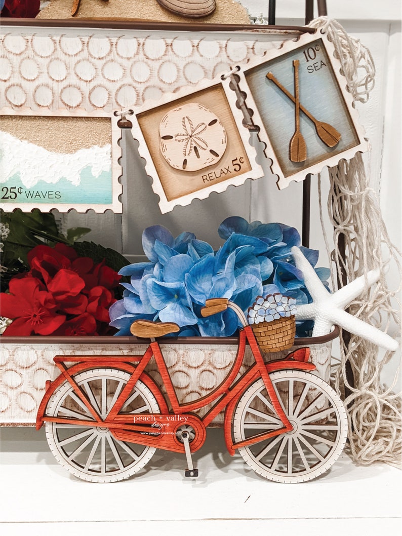 May include: A red wooden bicycle with a basket filled with blue flowers. The bicycle is sitting on a white surface with a brown wooden tray behind it. The tray has a variety of wooden signs with nautical themes, including a seashell, oars, and the words "Relax 5c" and "10c Sea".