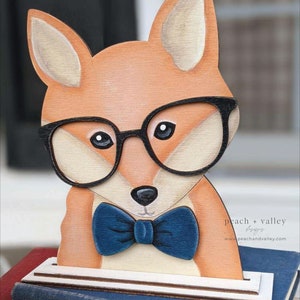 May include: A wooden fox figurine wearing black-rimmed glasses and a blue bow tie. The fox is painted in shades of orange, brown, and white. The figurine is standing on a stack of books.