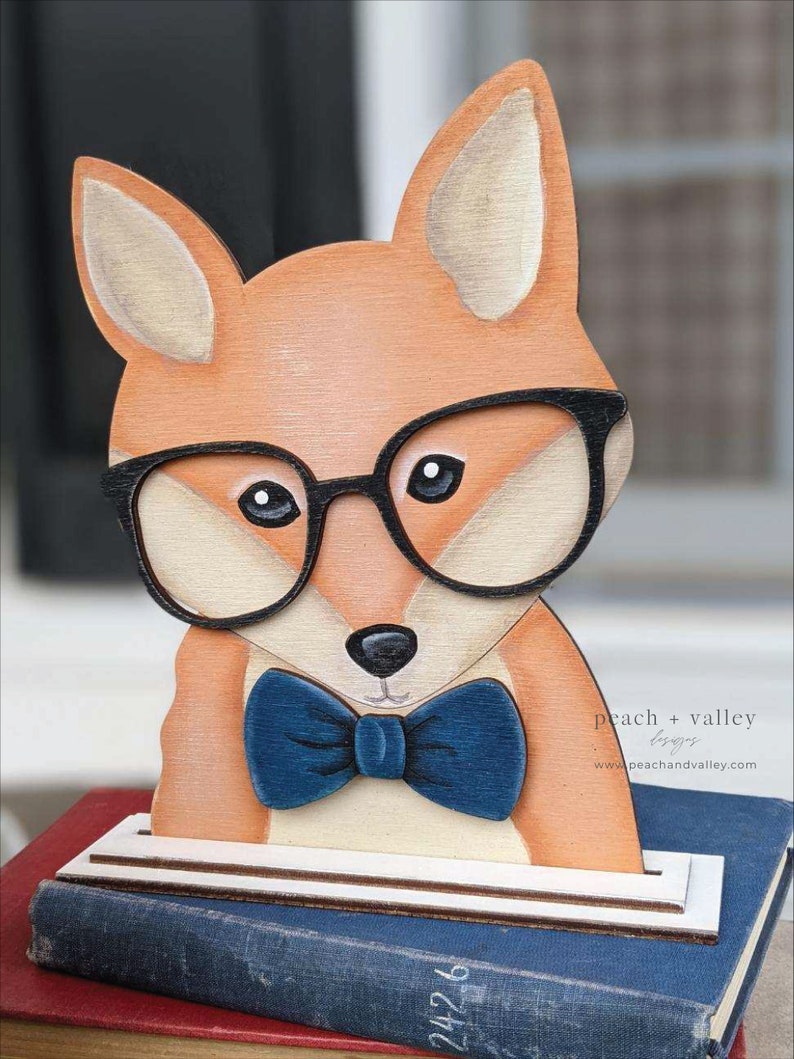 May include: A wooden fox figurine wearing black-rimmed glasses and a blue bow tie. The fox is painted in shades of orange, brown, and white. The figurine is standing on a stack of books.