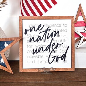 May include: A framed wooden sign with a white background and black text. The text reads "I pledge allegiance to the flag of the United States of America, and to the Republic for which it stands, one nation under God, indivisible, with liberty and justice for all." The sign is decorated with a red, white and blue star and a small American flag.