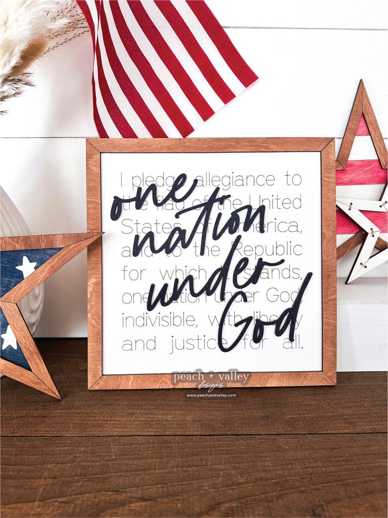 May include: A framed wooden sign with a white background and black text. The text reads "I pledge allegiance to the flag of the United States of America, and to the Republic for which it stands, one nation under God, indivisible, with liberty and justice for all." The sign is decorated with a red, white and blue star and a small American flag.