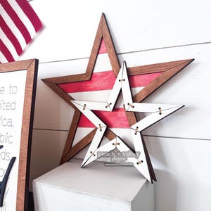May include: Two wooden stars, one red and white striped and one white, are layered on top of each other. The stars are decorated with brown stitching around the edges. The stars are on a white surface.