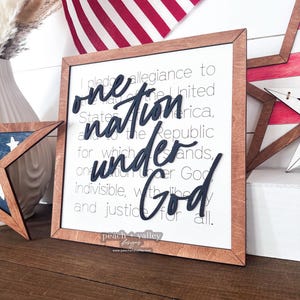 May include: A wooden sign with a white background and brown frame. The sign features the text "I pledge allegiance to the flag of the United States of America, and to the Republic for which it stands, one nation under God, indivisible, with liberty and justice for all." The words "one nation under God" are in black raised lettering.