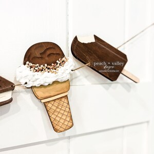 May include: Wooden ice cream cone and popsicle cutouts with a white background. The ice cream cone has a brown scoop of ice cream with white frosting and brown sprinkles. The popsicle is brown with a white bite taken out of the top. The text "peach + valley designs" is printed on the popsicle.