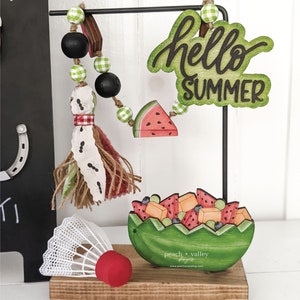 May include: A wooden sign with the text "hello summer" in green and black lettering. The sign is hanging from a black metal stand. A green and white gingham ribbon is attached to the stand. A wooden watermelon slice is hanging from the ribbon. A wooden watermelon bowl filled with fruit is on the stand. A white badminton birdie is on the stand. The stand is on a wooden base.