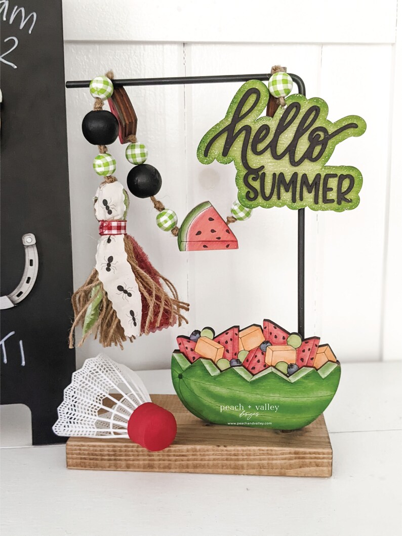 May include: A wooden sign with the text "hello summer" in green and black lettering. The sign is hanging from a black metal stand. A green and white gingham ribbon is attached to the stand. A wooden watermelon slice is hanging from the ribbon. A wooden watermelon bowl filled with fruit is on the stand. A white badminton birdie is on the stand. The stand is on a wooden base.