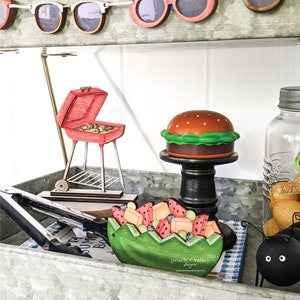 May include: A metal tray with a variety of summer-themed wooden decorations, including a watermelon fruit salad, a hamburger, a grill, and sunglasses. The tray is decorated with a blue and white checkered cloth and the text "peach + valley toys" is visible.