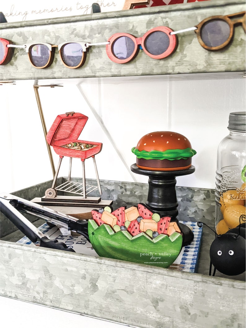 May include: A metal tray with a variety of summer-themed wooden decorations, including a watermelon fruit salad, a hamburger, a grill, and sunglasses. The tray is decorated with a blue and white checkered cloth and the text "peach + valley toys" is visible.