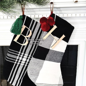 Op de afbeelding: Twee kerstsokken met zwart-witte patronen. De ene sok heeft een zwart-wit ruitpatroon met een groene kwast en een houten letter B. De andere heeft een zwart-wit blokpatroon met rode pompons en een houten letter H.