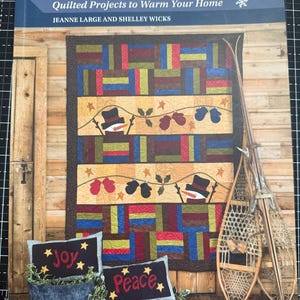 May include: A book cover titled "Here Comes Winter" featuring a colorful quilt design with snowman and mitten motifs. Decorative pillows with the words "Joy" and "Peace" are displayed in a metal container. Snowshoes and skis lean against a wooden wall.
