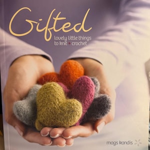 Op de afbeelding: De boekomslag van "Gifted" met de ondertitel "lovely little things to knit + crochet". De afbeelding toont handen die verschillende hartvormige, gebreide items in verschillende kleuren vasthouden, waaronder oranje, roze en groen.