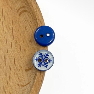 May include: Two round buttons with two holes each. One button is solid glossy blue, while the other is translucent white with a blue snowflake graphic printed on the surface. The buttons are positioned on a curved wooden surface.