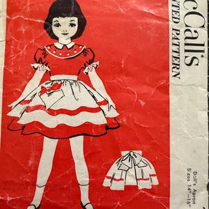 May include: A vintage McCall's sewing pattern for a doll's apron. The pattern is for sizes 14"-16" and features a red and white striped apron with a bow. The pattern is printed on a red background with the McCall's logo and the words "Printed Pattern" in white.