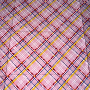 May include: Pink plaid fabric featuring a repeating pattern of red, yellow, blue, and white lines. The lines intersect to form a grid design. The fabric has a soft, textured appearance.