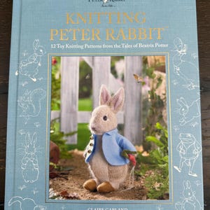 May include: A light blue hardcover book titled Knitting Peter Rabbit: 12 Toy Knitting Patterns from the Tales of Beatrix Potter by Claire Garland. The cover features a knitted Peter Rabbit figure in a blue jacket holding a carrot in a garden.