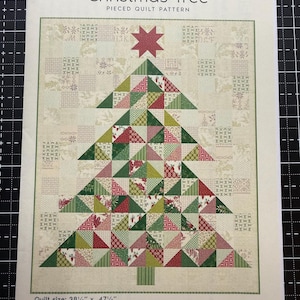 Könnte beinhalten: Ein Laundry Basket Quilts Christmas Tree Patchwork-Quiltmuster mit einem dreieckigen Baum aus roten, grünen und cremefarbenen Stoffquadraten. Das Design umfasst einen roten Stern als Spitze und einen grünen Stamm. Die Größe beträgt 97,8 x 120,6 cm.