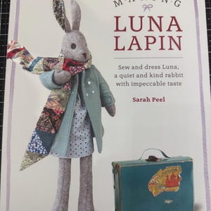 Op de afbeelding: Een boekomslag getiteld Making Luna Lapin door Sarah Peel met een grijze viltkonijnpop in een lichtblauwe jas, een jurk met stippen en een kleurrijke patchwork sjaal, met een kleine blauwe koffer en de tekst Includes 20 patterns.