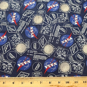 May include: Blue fabric with a repeating pattern of the NASA logo, a red and blue rocket, and a white circle with a moon-like surface. The text "INSPIRING THE WORLD THROUGH DISCOVERY" is repeated throughout the fabric.